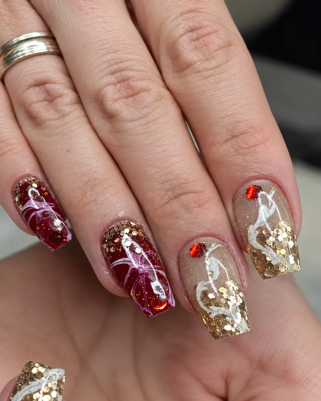 Nahaufnahme der Nägel mit Goldglitzer, weißen Schnörkeln und roten Steinchen